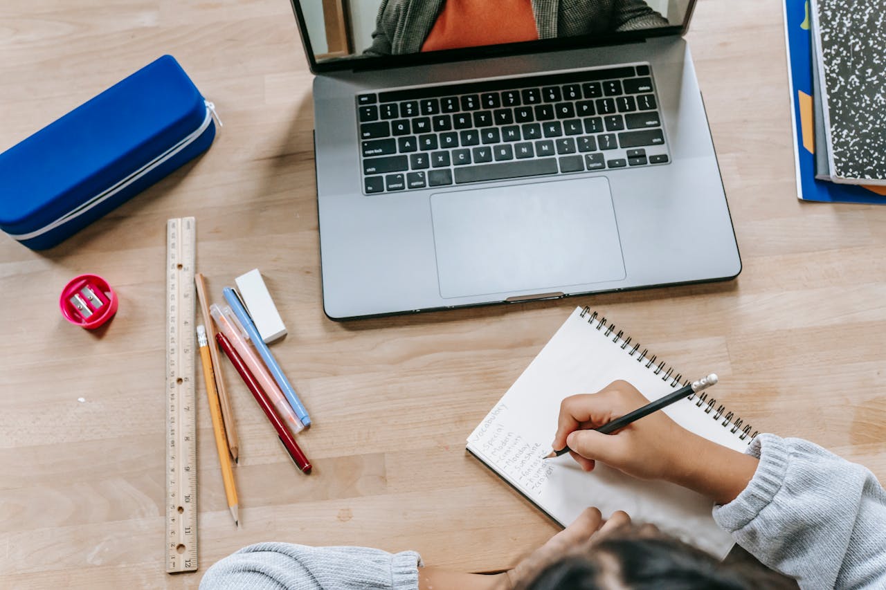 Top view of crop anonymous smart pupil writing information in notepad while studying online with laptop at table with stationery