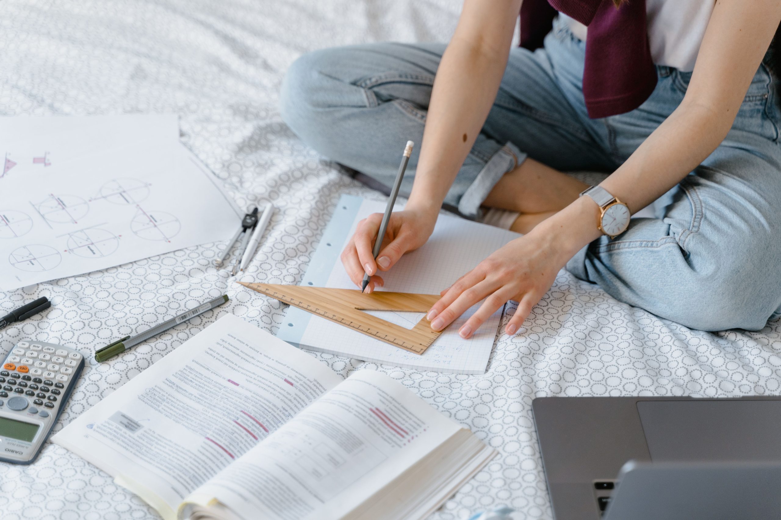 Student uses geometry tools and laptop to study math concepts while sitting on bed.